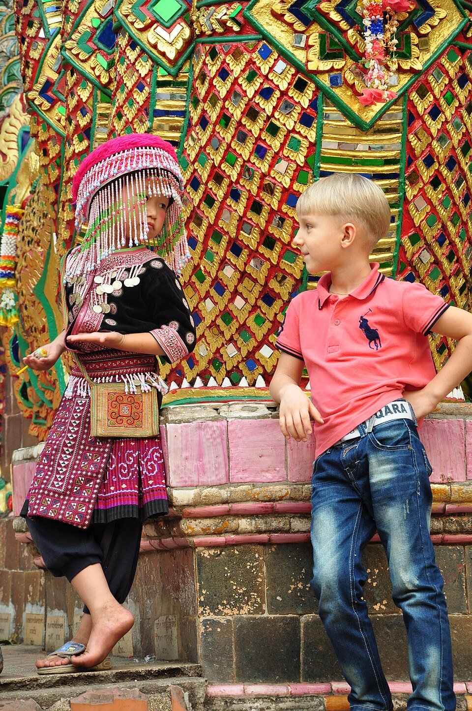 A girl in traditional Hmong attire interacts with a young tourist at a temple in Thailand. (Wikimedia)